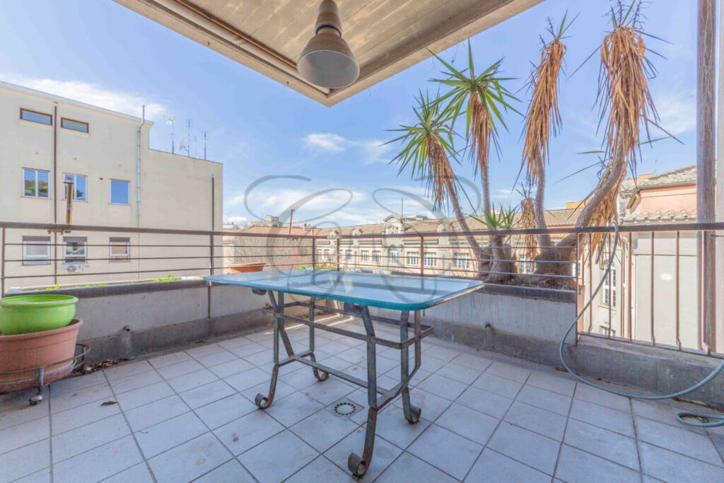 Terrazzo semicoperto con un tavolo e sedie. La vista si apre su un cielo azzurro con nuvole e tetti di Roma.