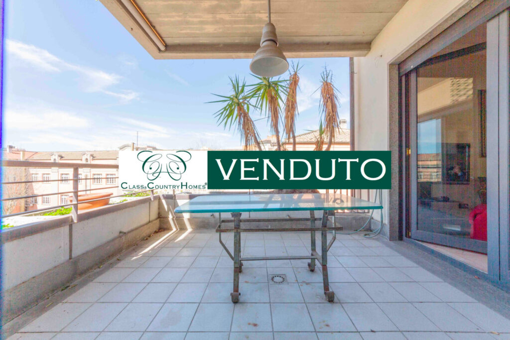 Un terrazzo arredato con un tavolo e una rete da tennis, con il cartello "VENDUTO" in evidenza. Lo sfondo mostra il cielo e i tetti di Roma.